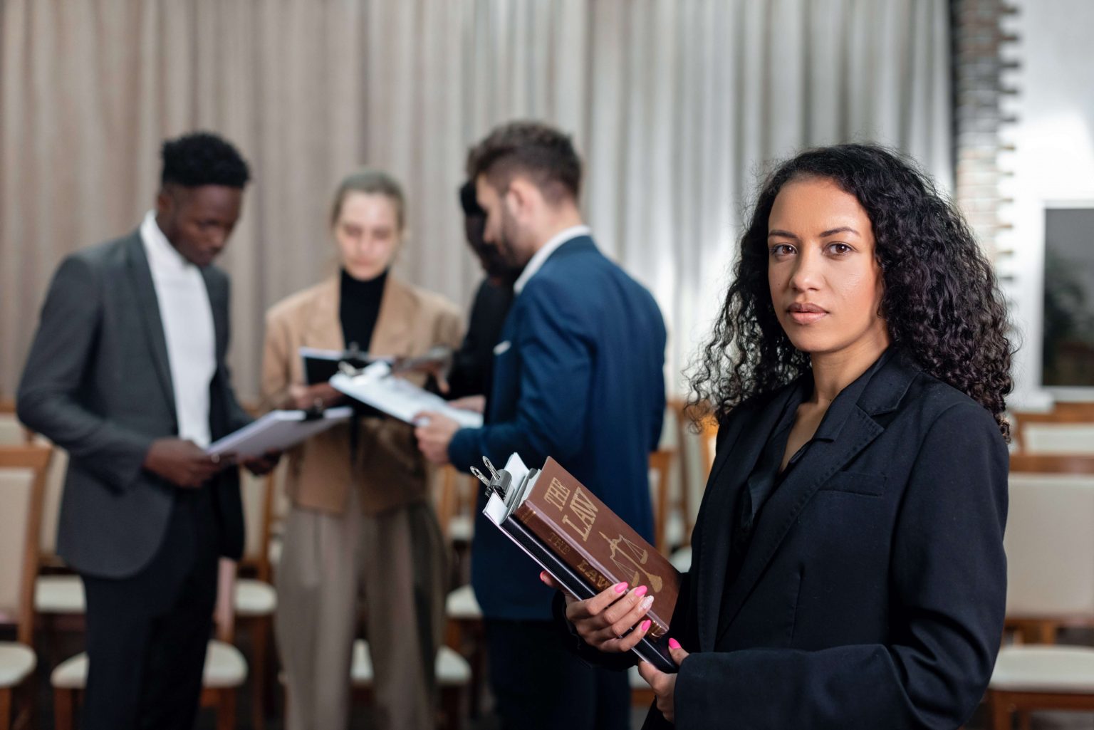 A woman in a suit holding books and papers stands in the foreground, while four people have a discussion in the background of a conference room.