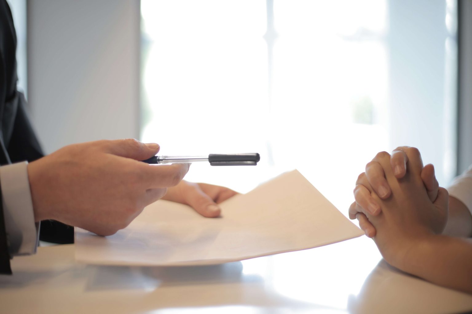 Two people sit at a table; one person holds documents and offers a pen, while the other has hands clasped, apparently in a discussion or agreement.