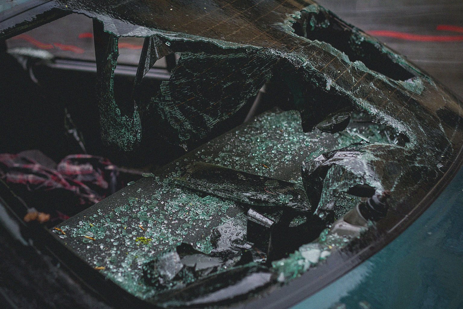 A car windshield shattered inward with large broken glass pieces covering the dashboard and front seats.