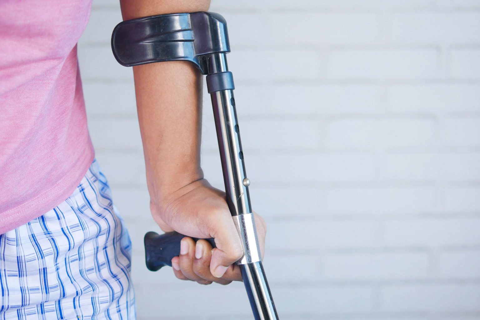 Person wearing a pink shirt and plaid shorts holding a forearm crutch against a light-colored brick wall background.