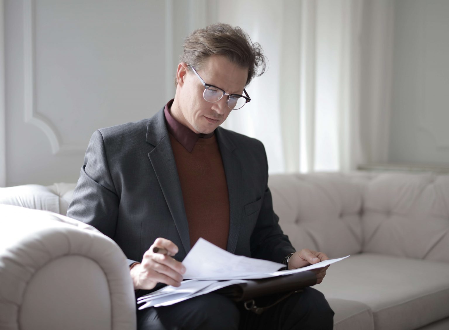A man in a suit and glasses sits on a light-colored sofa, reviewing documents with a focused expression.