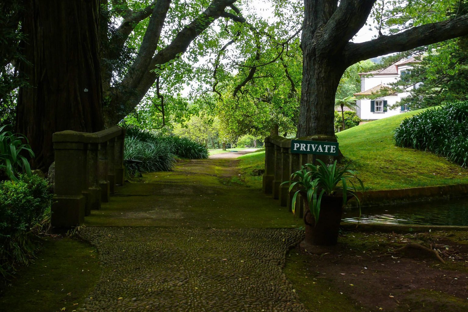 Stone pathway with moss leads over a small bridge toward a shady garden; a "PRIVATE" sign is posted beside the bridge, and a white house is visible in the background.