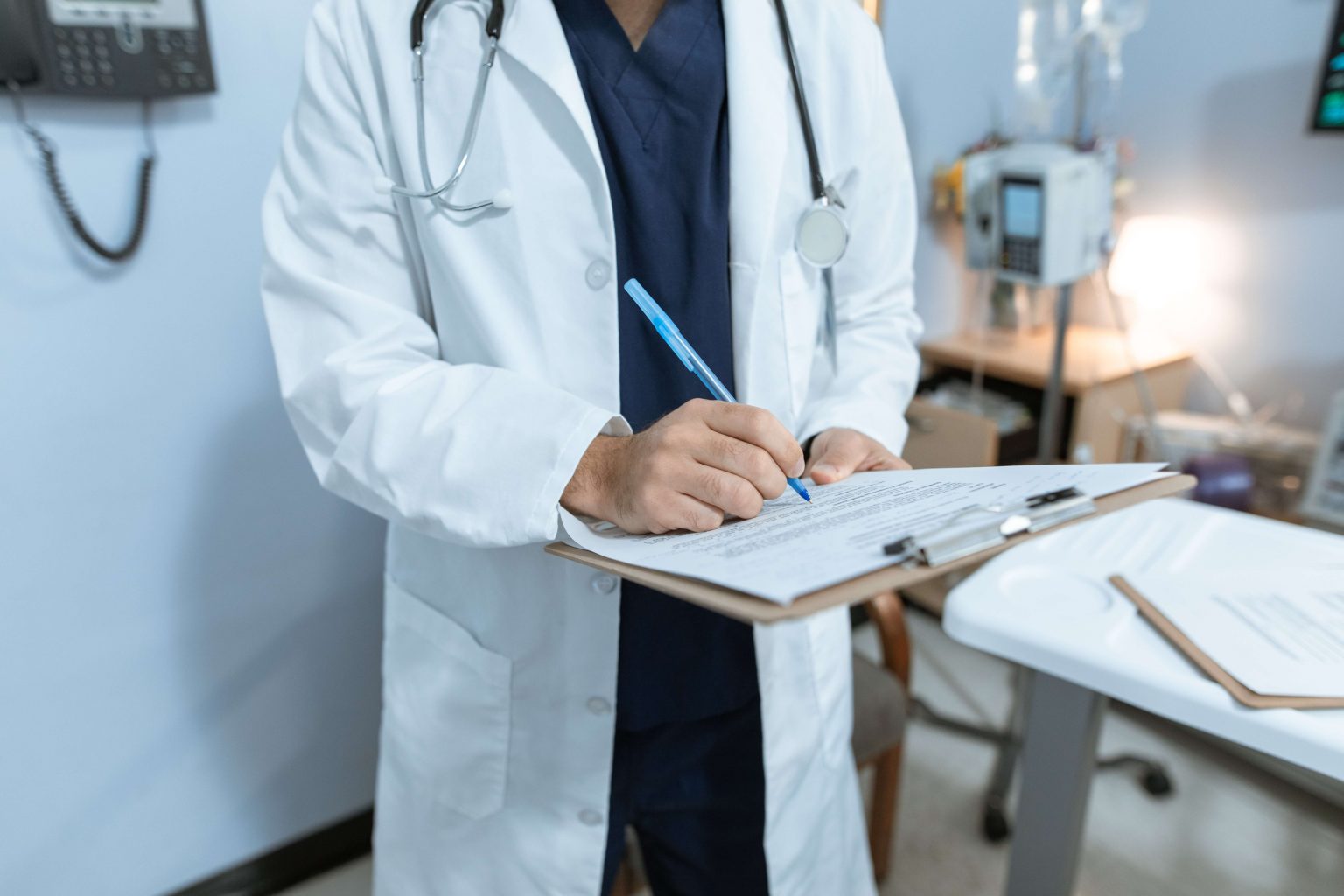 A doctor in a white coat and stethoscope writes on a clipboard in a medical office.