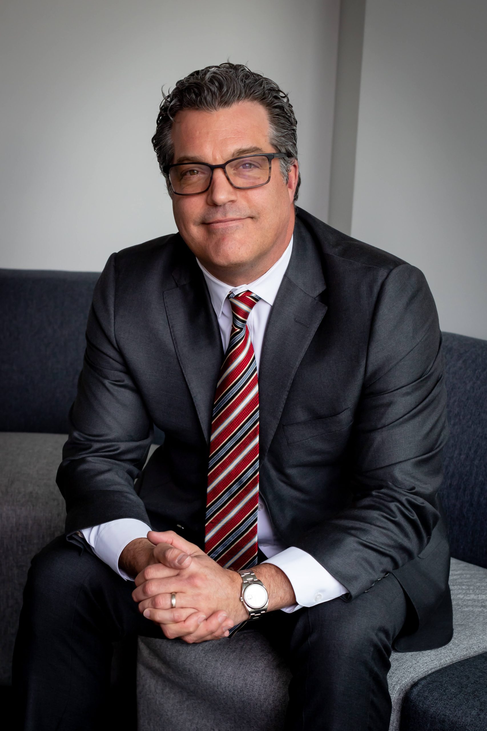 A man in a dark suit and striped tie sits on a gray couch with his hands clasped, looking at the camera and smiling slightly.
