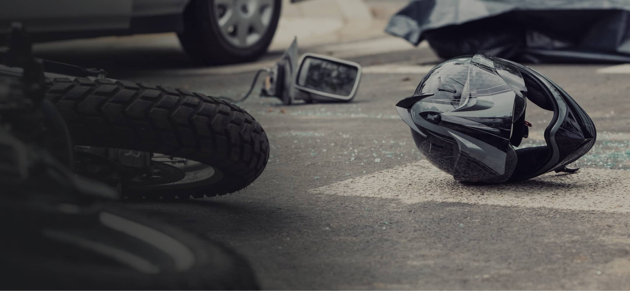 A black motorcycle helmet lies on the asphalt next to a fallen motorcycle and broken mirror, suggesting a recent accident scene.