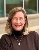 A woman with shoulder-length blonde hair wearing a dark turtleneck sweater and a gold necklace smiles in front of a blurred building background.