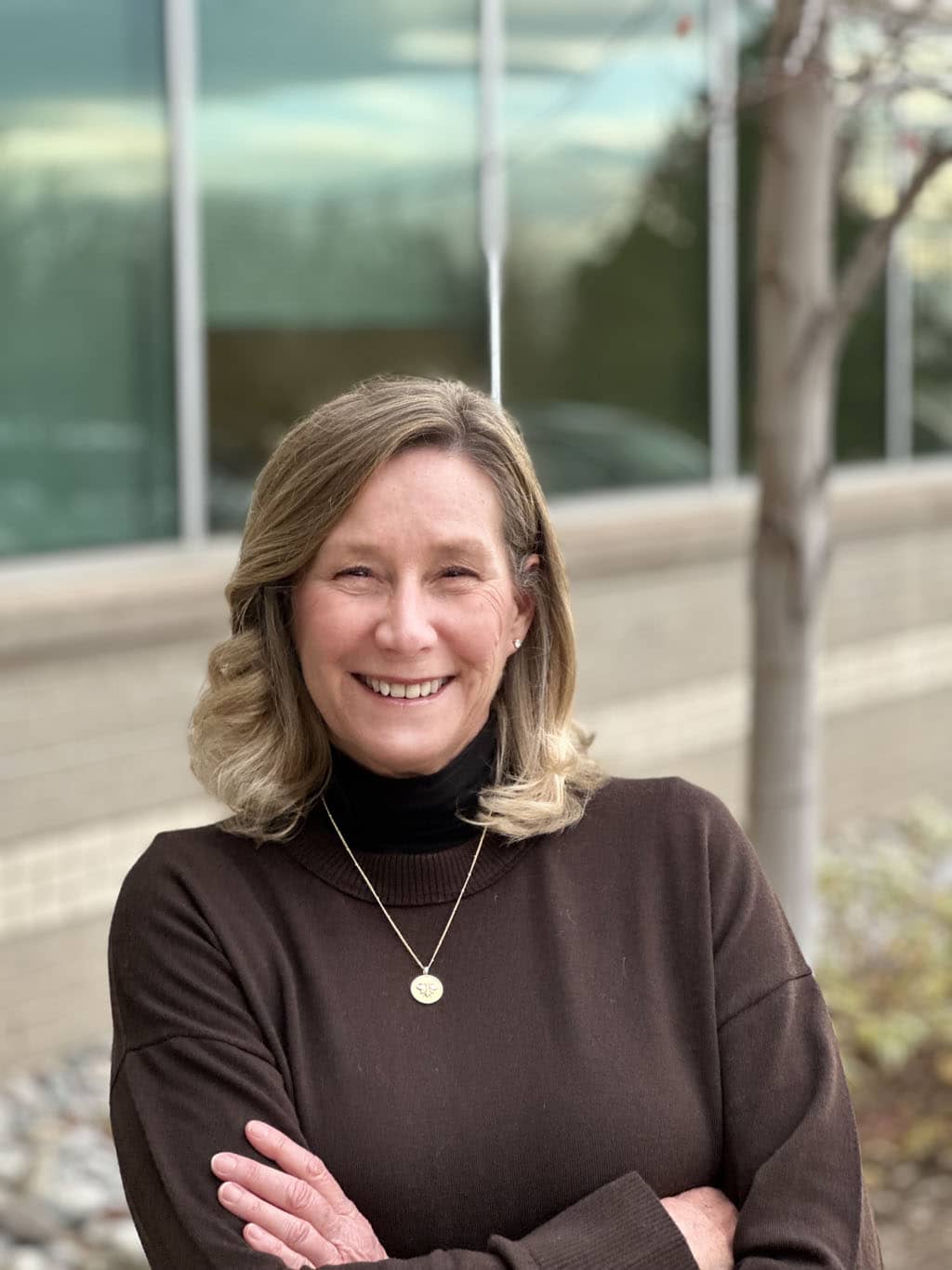 A woman with shoulder-length blonde hair, wearing a brown sweater and gold necklace, stands outdoors with her arms crossed, smiling in front of a building with large windows.