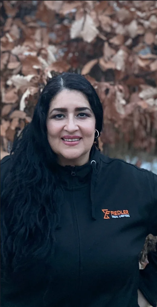 A woman with long black hair smiles at the camera, wearing a black jacket with an orange "REDLER Stahl Logistik" logo. Brown leaves are visible in the blurred background.
