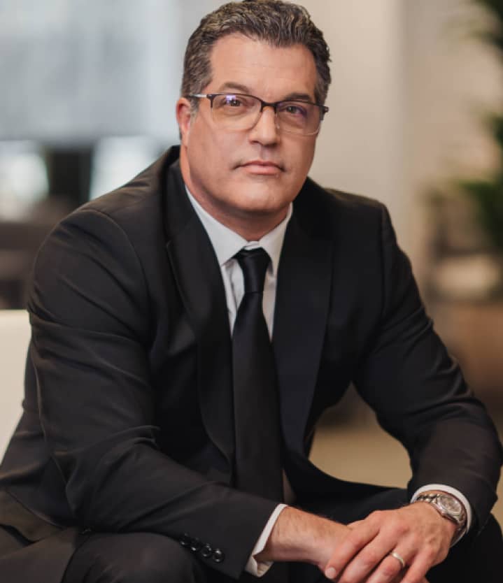A man in a black suit and tie, wearing glasses, sits indoors with a neutral expression.