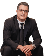 A man wearing a dark suit, tie, and glasses is seated, looking at the camera with a neutral expression.