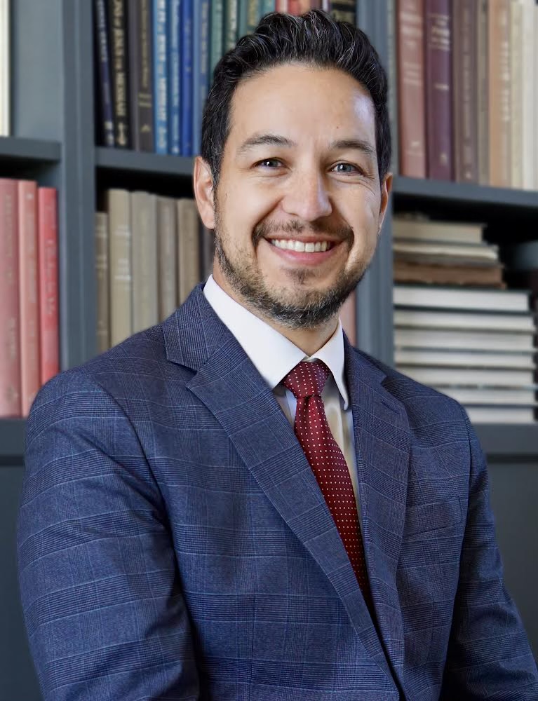 A man in a blue checked suit and red tie smiles while seated in front of shelves filled with books.