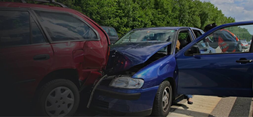 A blue car with a crumpled hood has rear-ended a red SUV on a road; people stand nearby, and trees line the background.