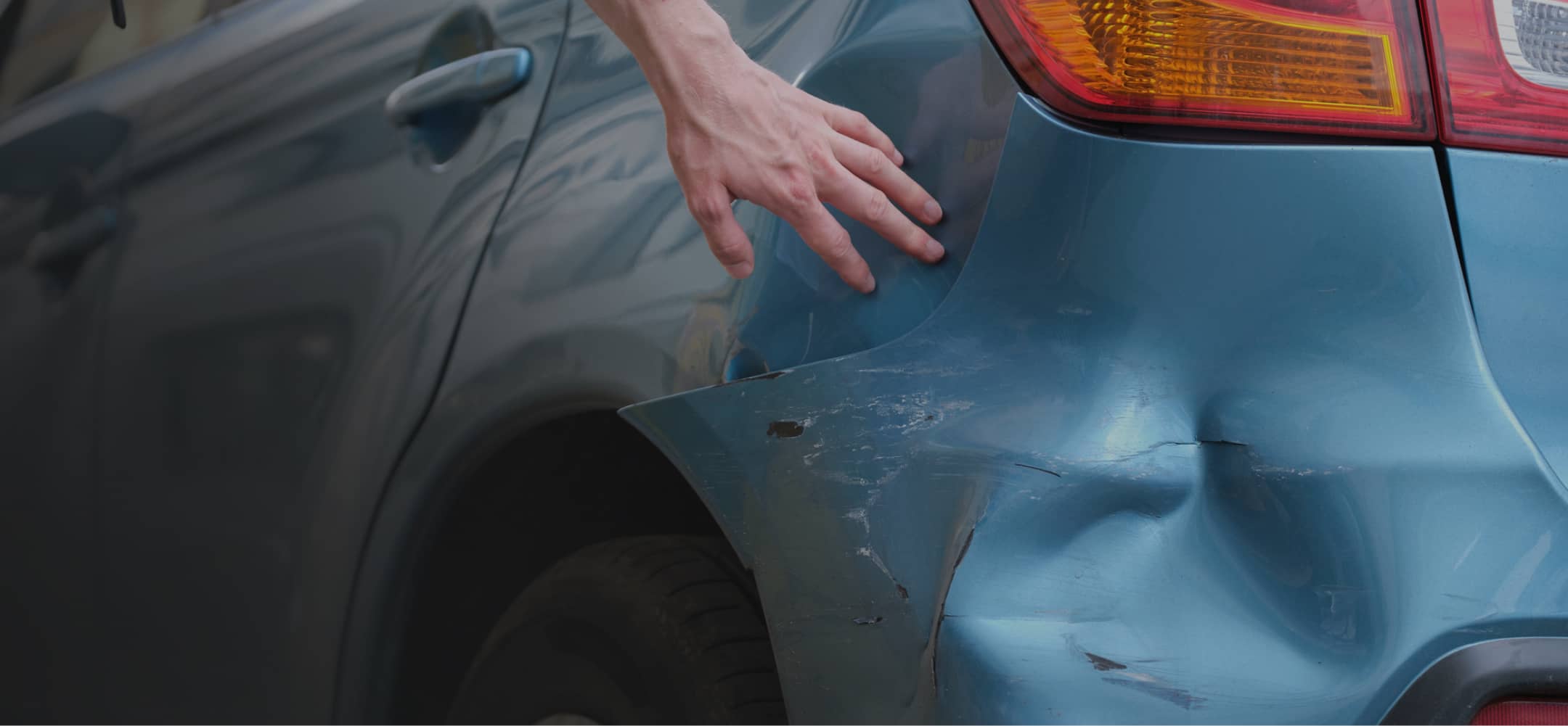 A hand points to a dent and scratches on the rear bumper of a blue car near the right tail light.