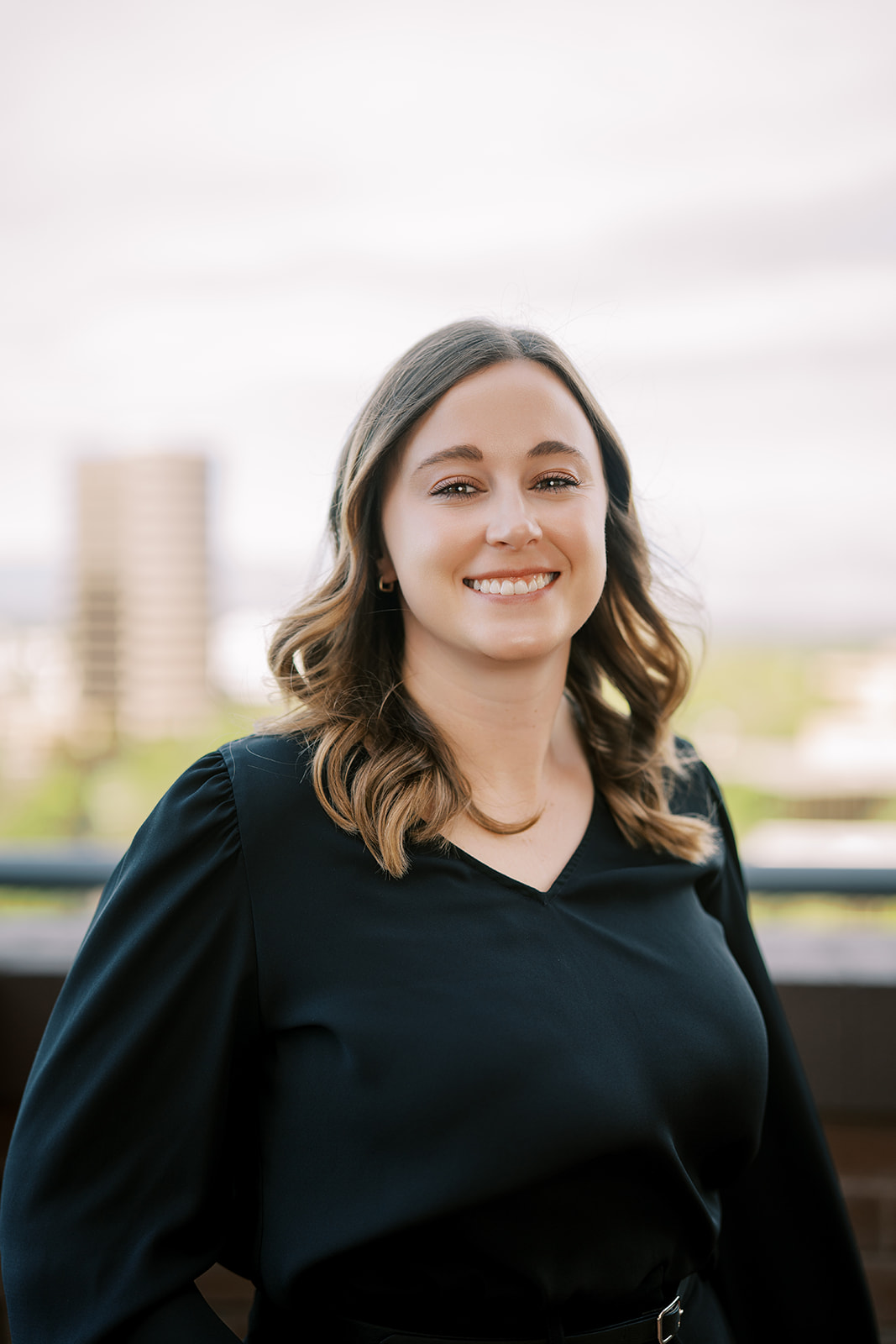Woman with shoulder-length brown hair wearing a black blouse, standing outside with a blurred cityscape in the background. She is smiling and facing the camera.