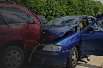 A blue car with a crumpled hood has rear-ended a red SUV on a road; people stand nearby, and trees line the background.