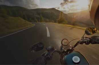 A person rides a motorcycle on a winding mountain road at sunset, with trees and hills visible in the background.
