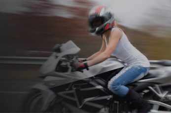 A person wearing a helmet, white tank top, and jeans is riding a black sports motorcycle on a road with a blurred background.