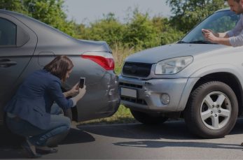 A woman and a man take photos of the damage after a car accident involving a gray sedan and a silver SUV on a sunny day.