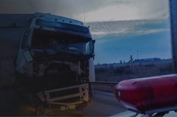 A damaged truck with a missing front hood is stopped on the side of a road near a police vehicle at dusk.